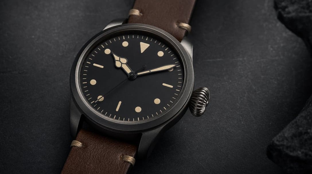 Large minimalist pilot’s watch with a clean black dial, oversized onion crown, and brown leather strap on dark slate