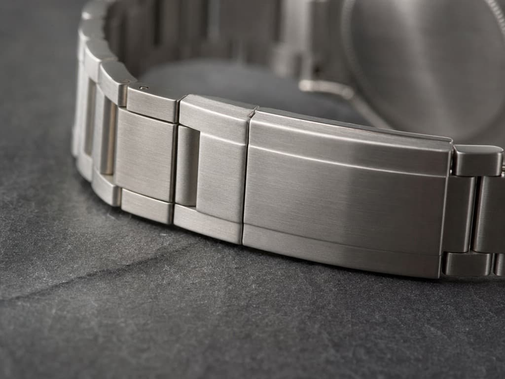 Macro view of a stainless steel watch bracelet showing solid links and a folding clasp closed flush against dark slate
