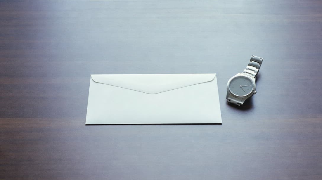 A single sealed white envelope alone at the centre of an otherwise empty desk beside a stainless-steel watch